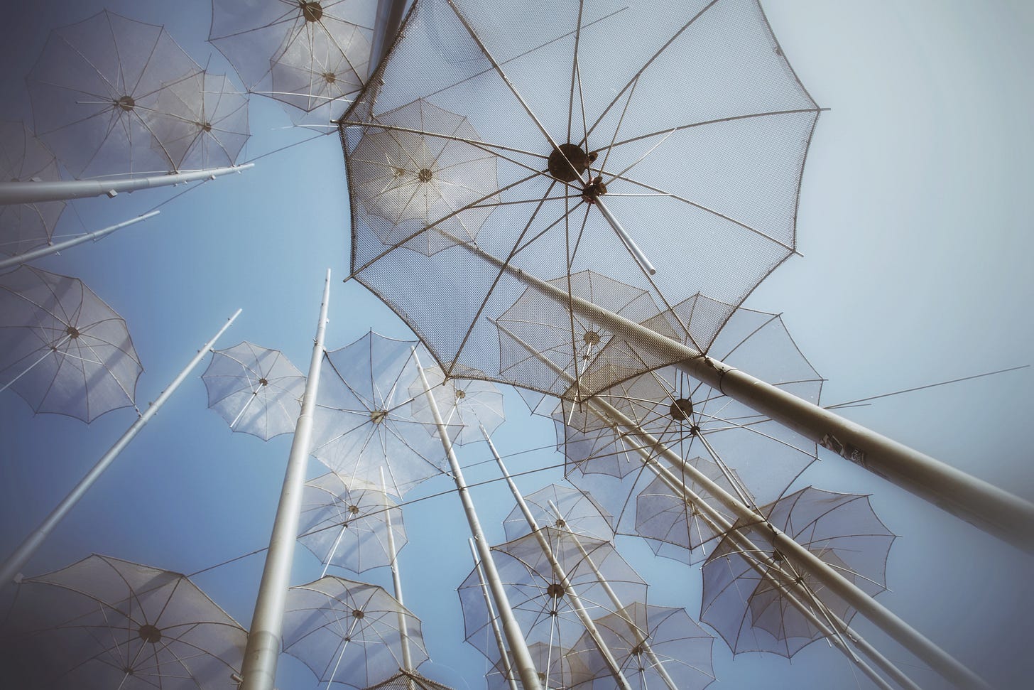 A photograph taken from below a public art installation of tall metal poles topped with translucent umbrellas against a pale blue sky. The umbrellas overlap and create a delicate, geometric pattern of lines and light.