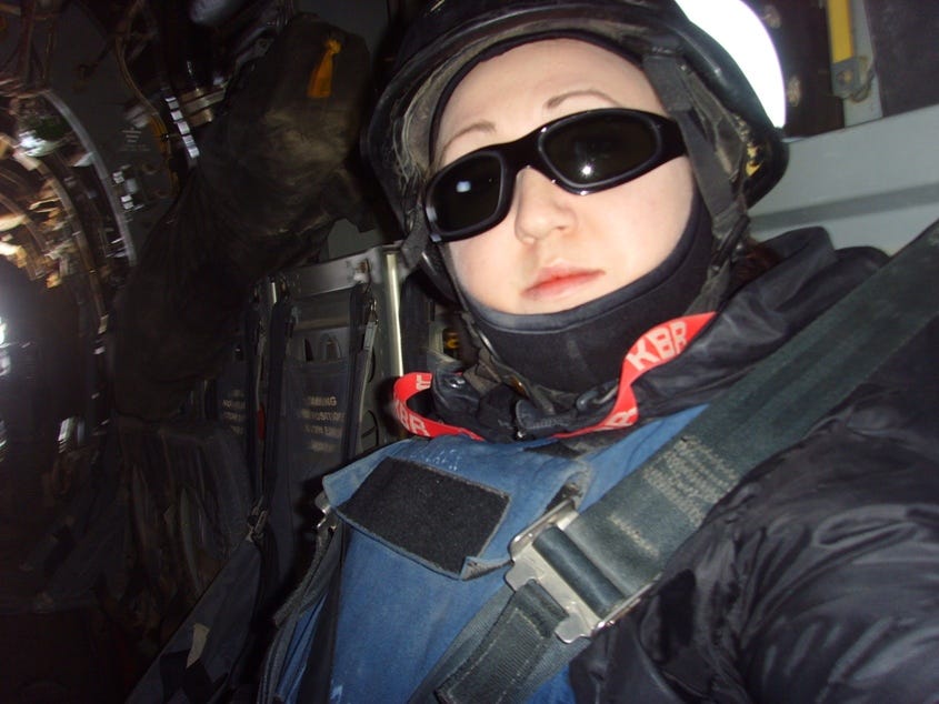 A photo of the author, a woman, wearing cold weather gear with a Kevlar vest and helmet, while riding in the back of an Osprey flight vehicle. A bright red KBR lanyard is visible and she is wearing protective goggles.