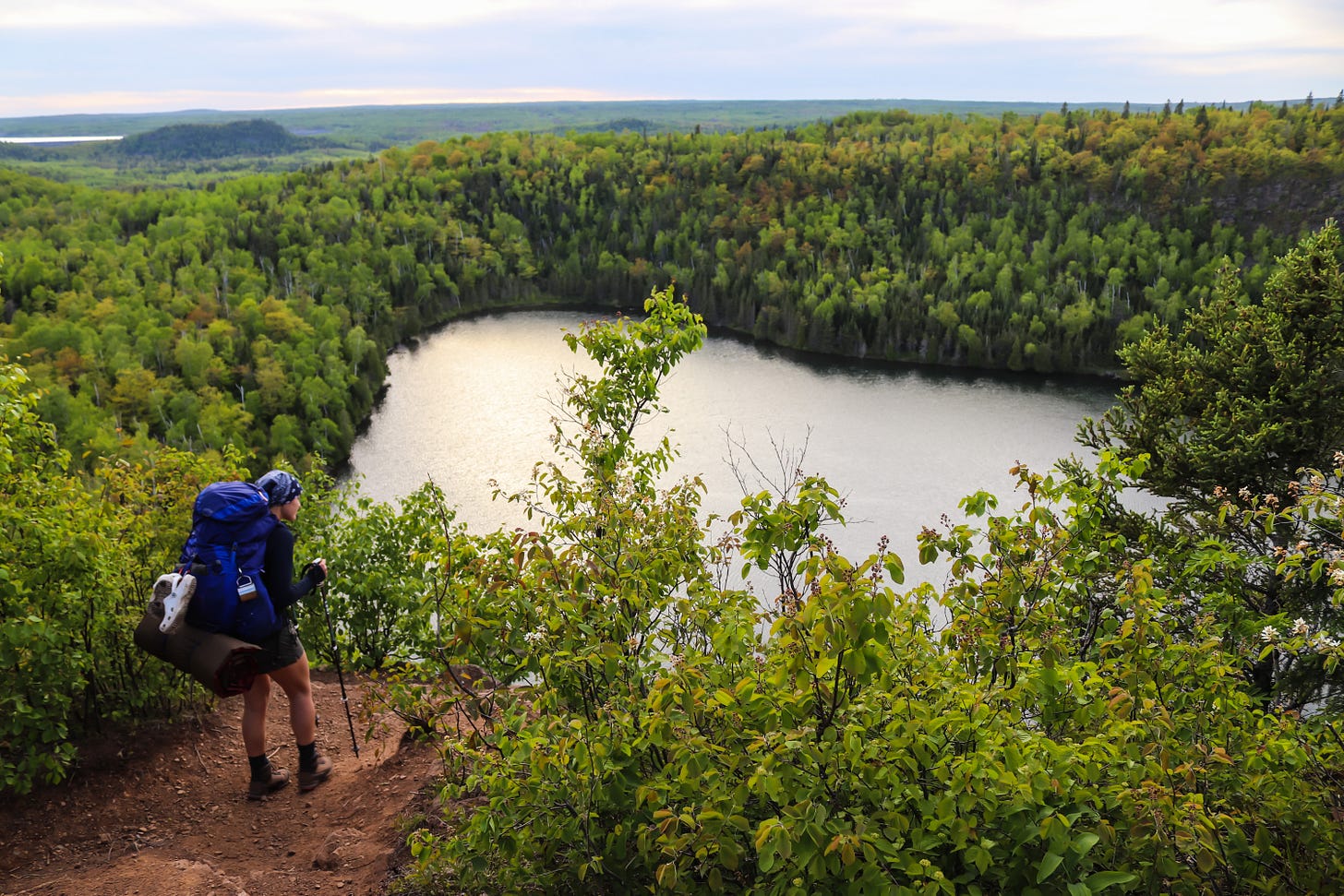 Picture shows Lisa hiking with a backpack with a gorgeous lake view on a cliff with green trees