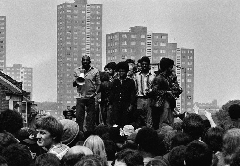 Black and white photo of a street protest from the 1980s in London. Darcus Howe is speaking to the assembled crowd through a megaphone on elevated platform with other speakers next to him. Grey concrete council flats are visible far in the background.