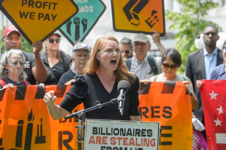 A woman speaks outdoors in front of a microphone as several peopple holding a banner stand behind her. A woman speaks outdoors in front of a microphone as several peopple holding a banner stand behind her.