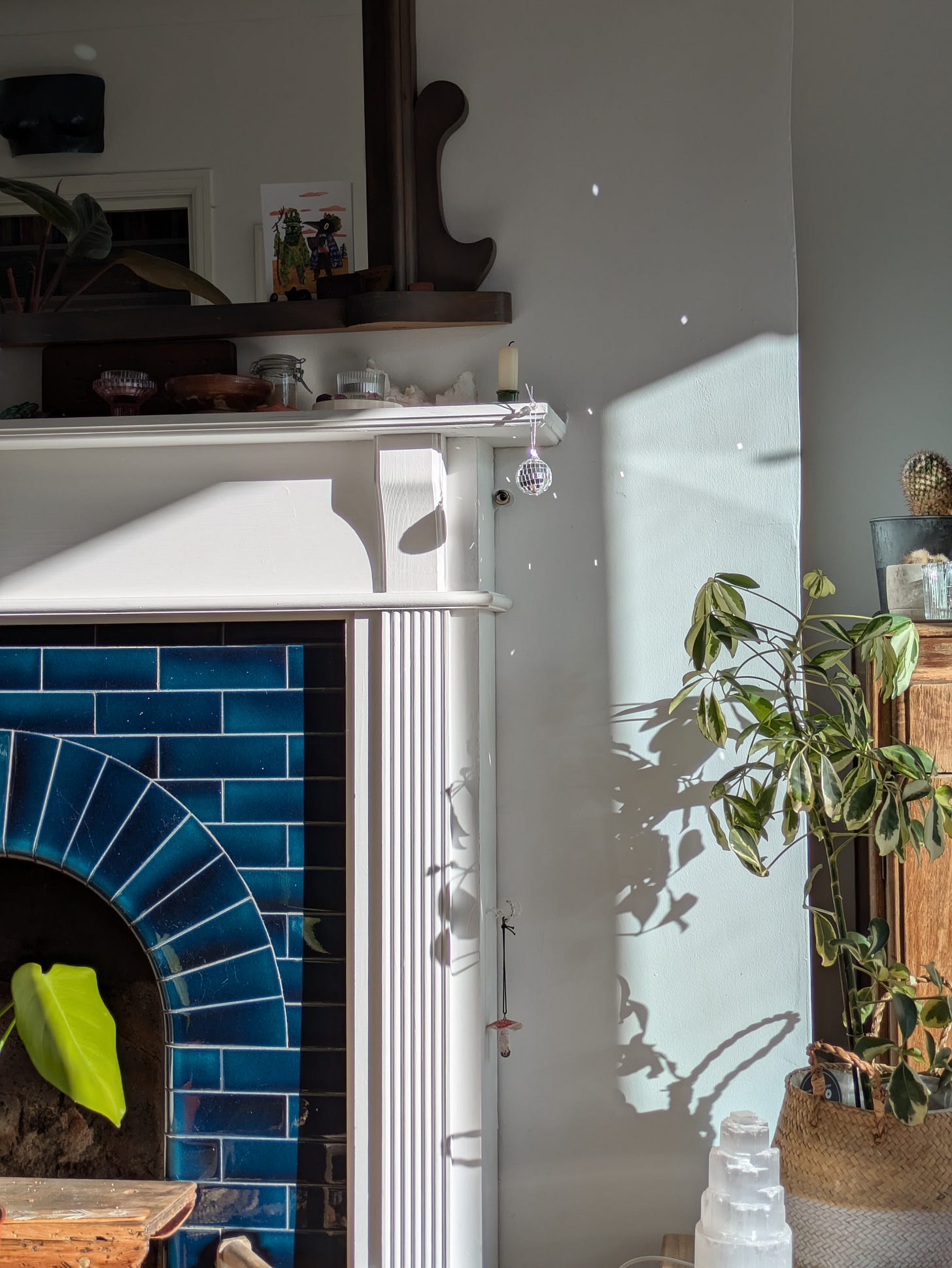 one side/corner of the mantelpiece and fireplace in Grace's living room, navy/teal tiles and white pillar on a white wall, plants decorating the space and a tiny disco ball glimmering in the sunlight