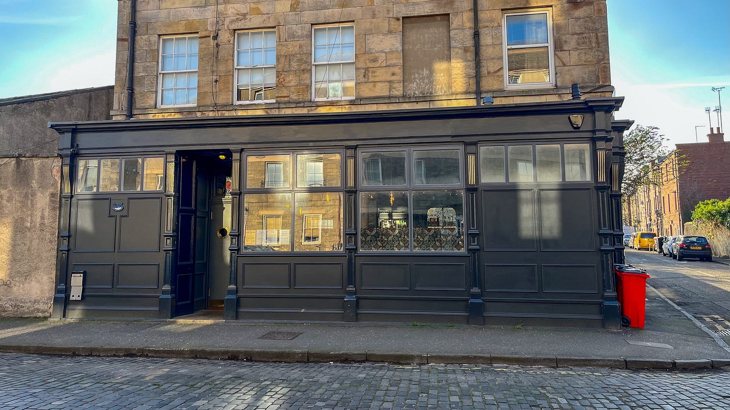The black exterior of a pub, on the corner of the street on the ground floor of a tenement building.