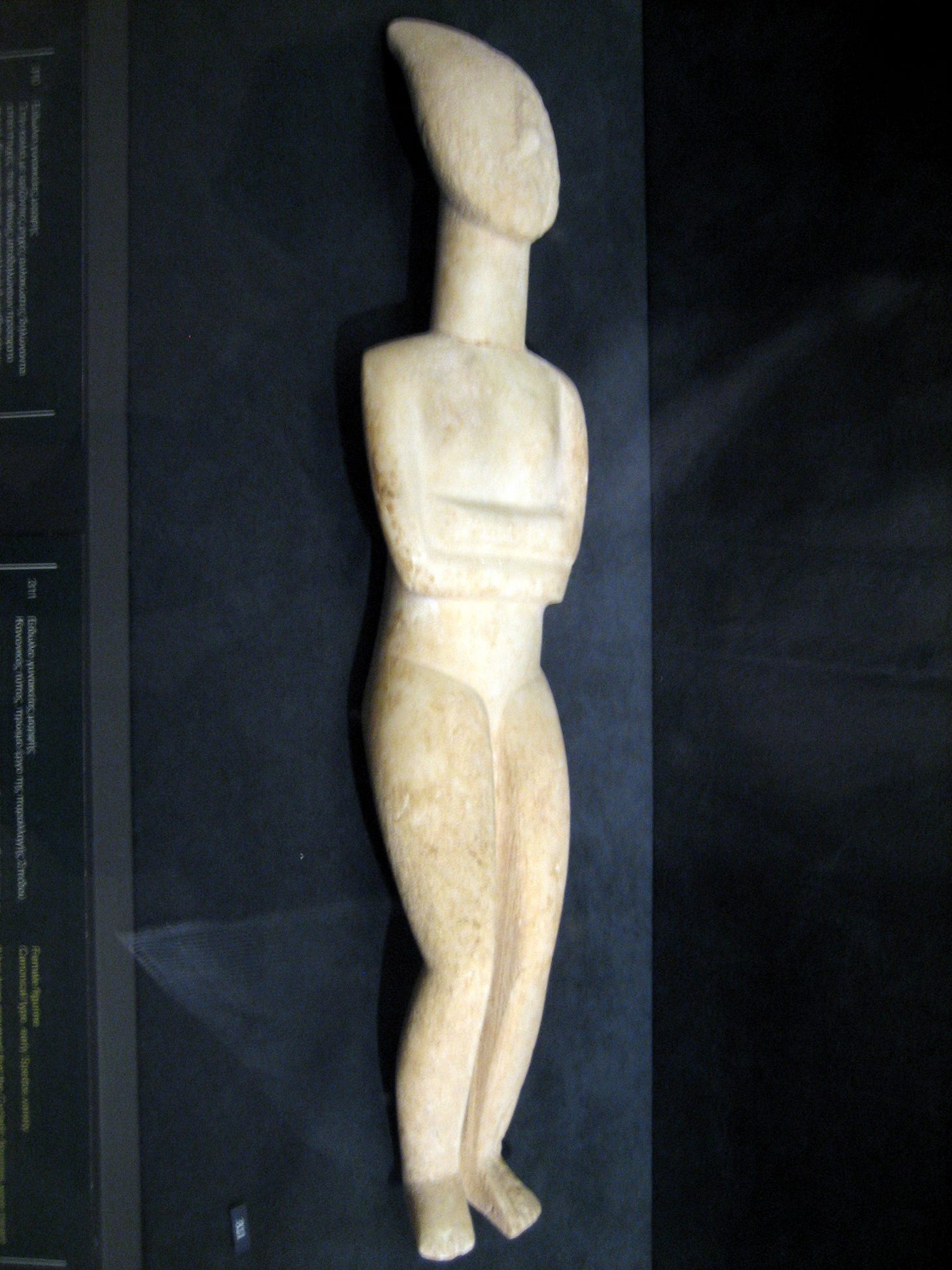 White marble Cycladic figurine viewed from the side, showing how the legs are bent, the head is tilted back, and the feet are held at an angle.