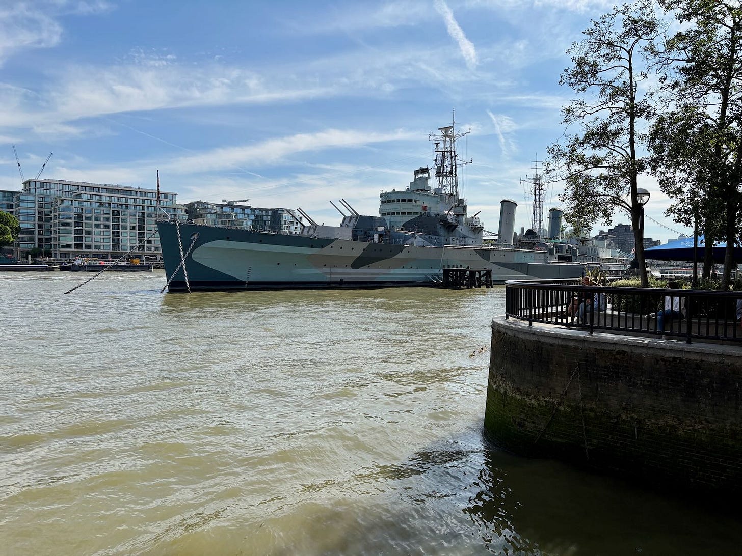 HMS Belfast, a large grey World War II-era Royal Navy warship, moored on the River Thames in London, with the city skyline in the background.