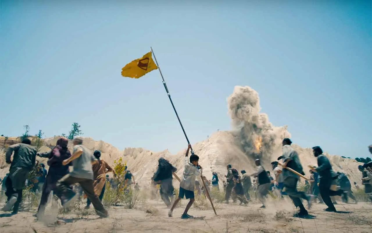 A young boy raising a yellow flag with the red Superman symbol on it while people in a desert are running around with explosions going off around them