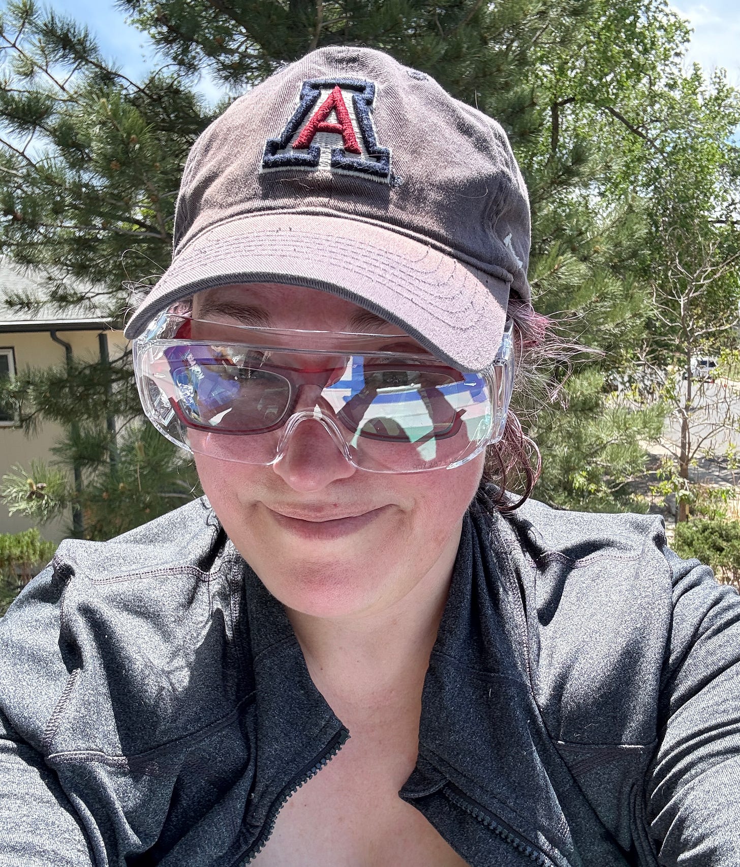 Me wearing a dark University of Arizona baseball cap, reflective safety goggles over regular glasses, and a zip-up athletic jacket smiles at the camera. She is outdoors on a sunny day, with pine trees and a building in the background.