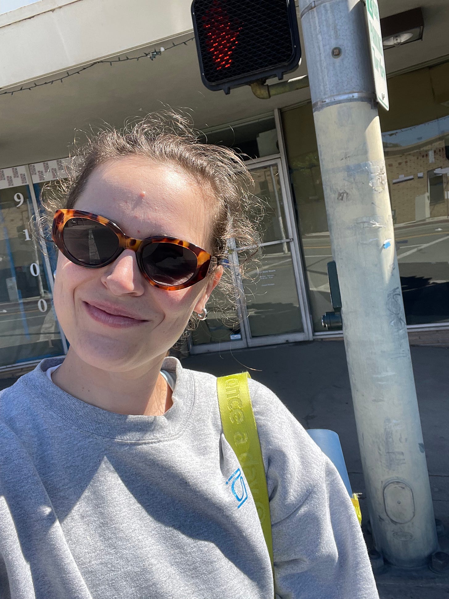 Woman at a crosswalk smiling at the camera wearing a gray sweatshirt and sunglasses and carrying a yoga mat.