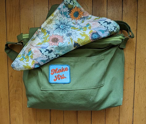 Five photos of the Mythical Messenger Bag. In the first, I (a white lady, aged 52) am standing in the hallway wearing the bag. The other photos are of the bag on its own. The bag is olive green canvas with an adjustable strap. There is hot pink decorative stitching. There is an exterior welt pocket on the back with a hot pink zipper. There is a pocket on the front with a blue square patch that says "Make Art" in a red cursive font. The inner lining fabric is floral -- blue, green, coral line drawn flowers on a white background. Inside the bag, there are two pockets. One is a zippered pocket with a hot pink zipper. The other is a divided pocket with slots for pens, a phone, and something else roughly cell-phone-sized.