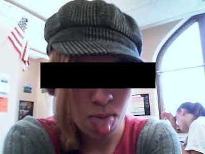 Grainy flip-phone photo of a blonde teen in a newsboy cap, sticking her tongue out at the camera. She is in an American high school classroom, the flag hanging in the background. Another student studies with her head down nearby. The main subject has been censored with a black bar covering her eyes.