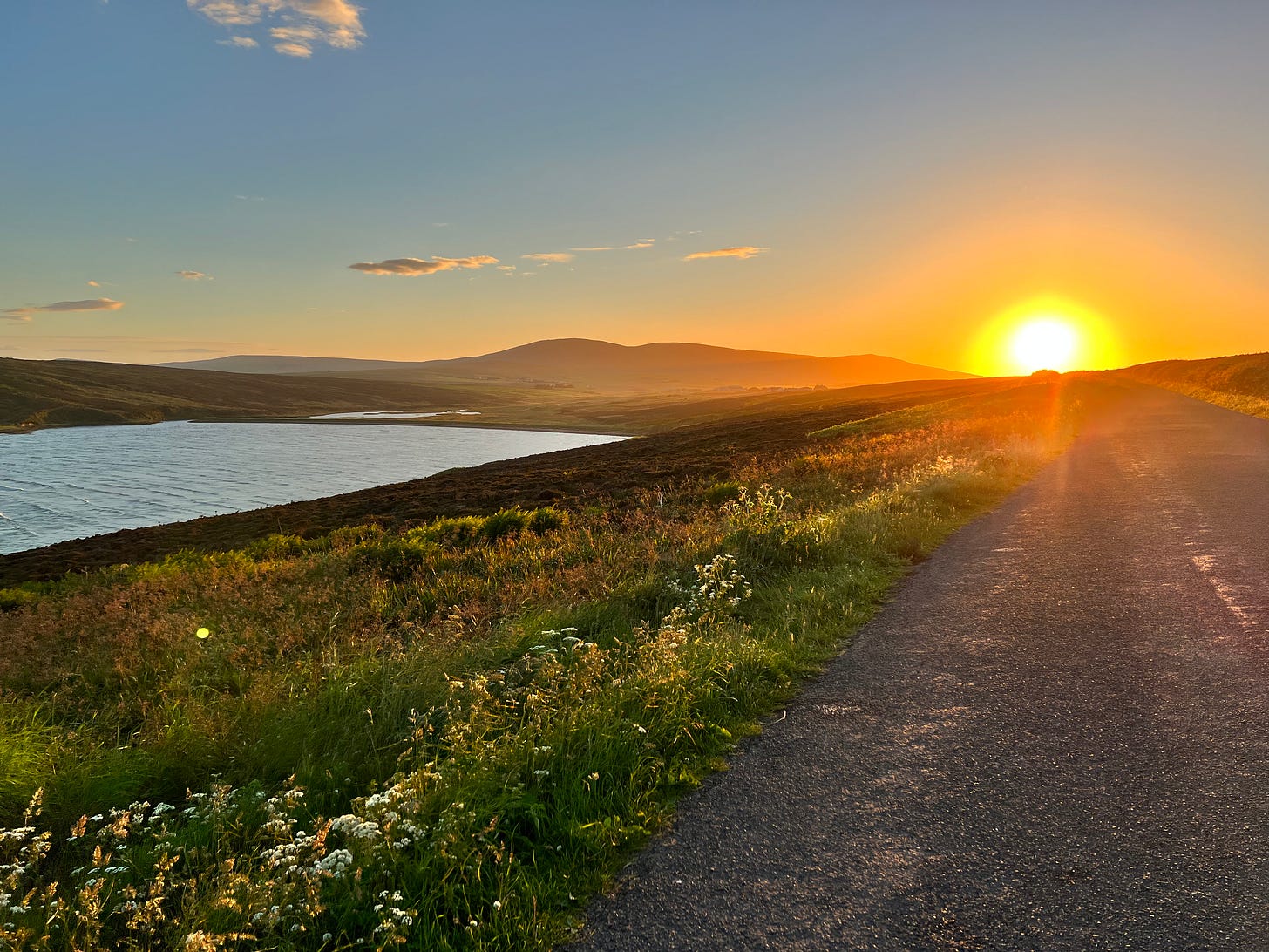 a long tarmac and gravel road stretching toward a setting sun. to the left is a bank of brightly lit grass and heather and to the right of that a stretch of water. I can't actually remember exactly where this is but it might be near the Ness of Brodgar