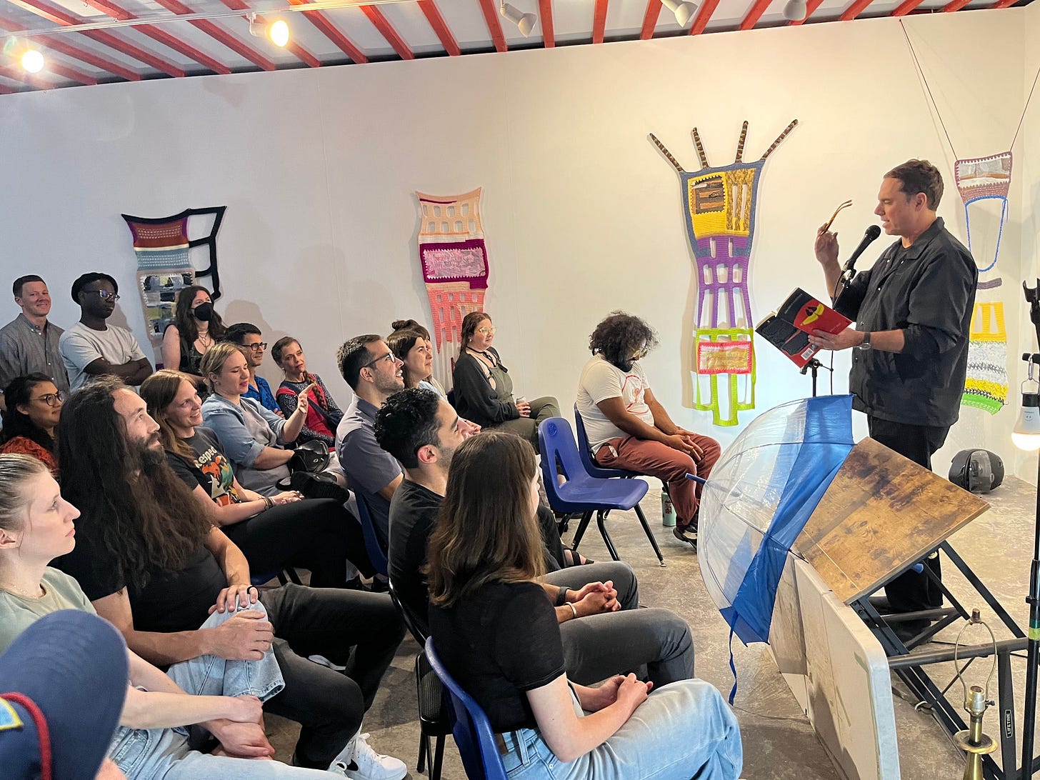 Derrick stands in front of a microphone, holding his book and performing. The audience watches him. A table, lamp, umbrella, and other objects are stacked together next to him. 