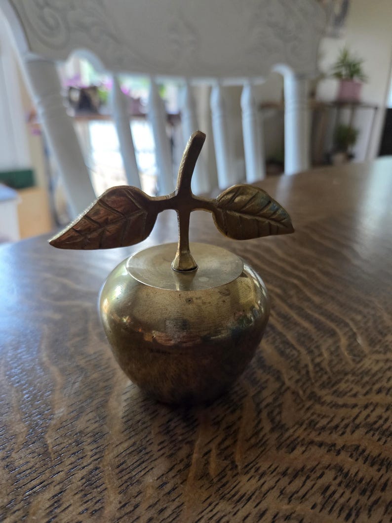 May include: A brass apple-shaped decorative object. The apple has a round body with a stem and two leaves. The object is made of shiny, golden-colored metal and sits on a wooden surface. The leaves are detailed with veins, and the stem is a simple, straight line.