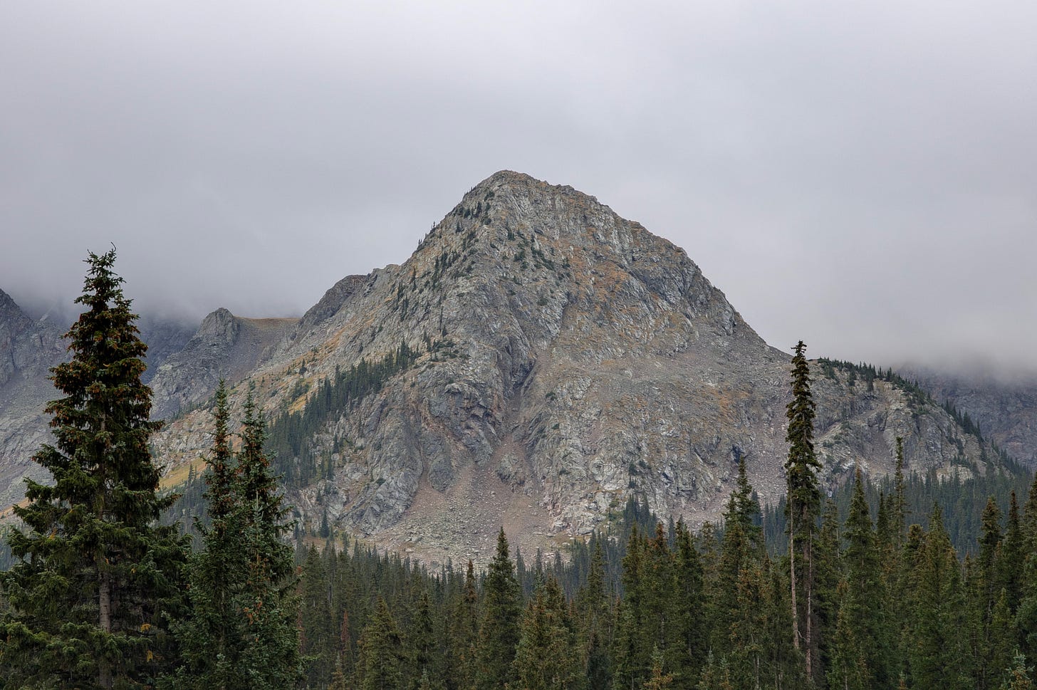 Rocky mountain peak rising above dense evergreen forest under low clouds, illustrating rugged alpine terrain and elevation challenges.