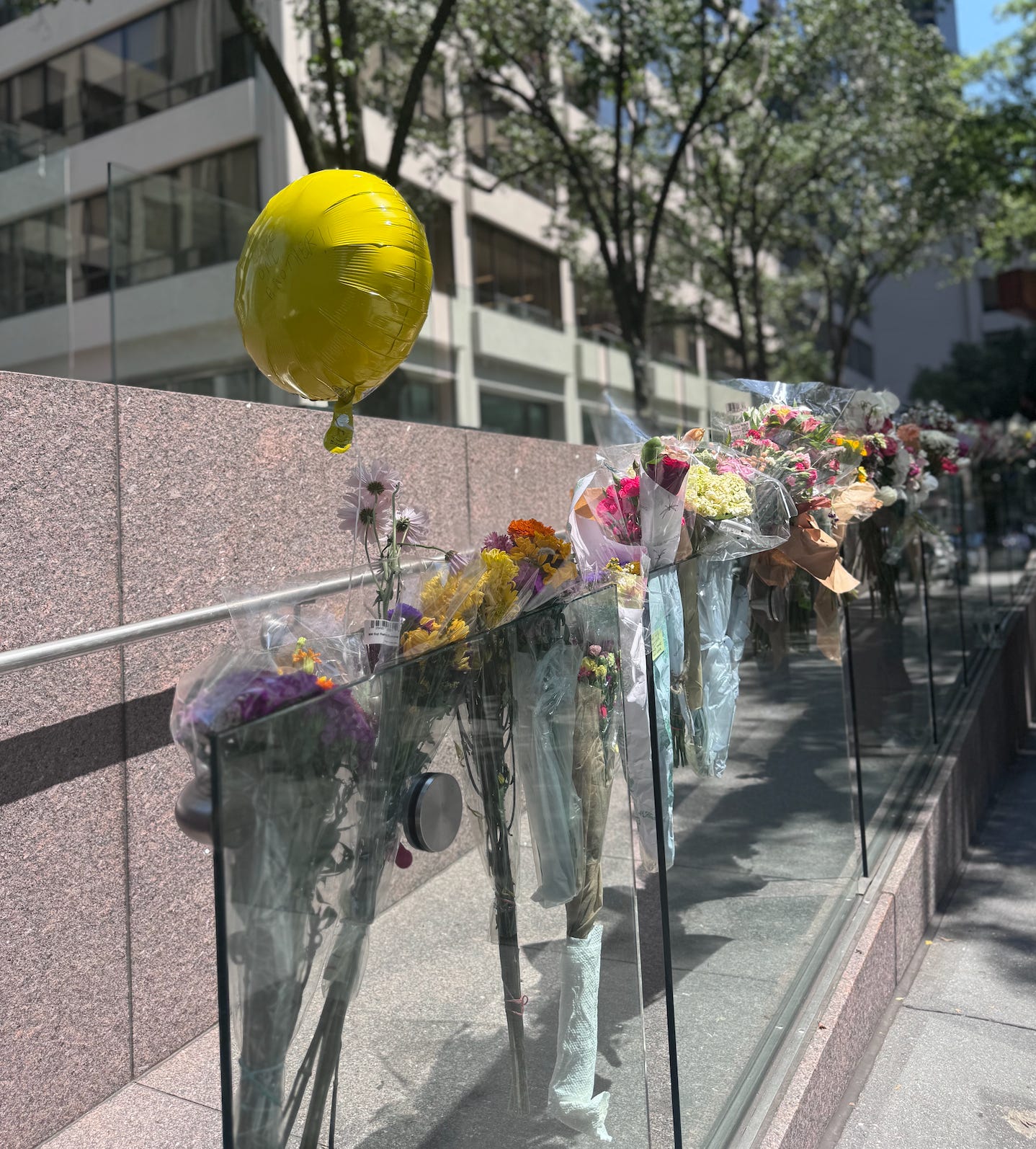 Bouquets of flowers have been secured onto a glass railing of a building's wheelchair ramp. A bright yellow helium balloon is tied to one of the bouquets.