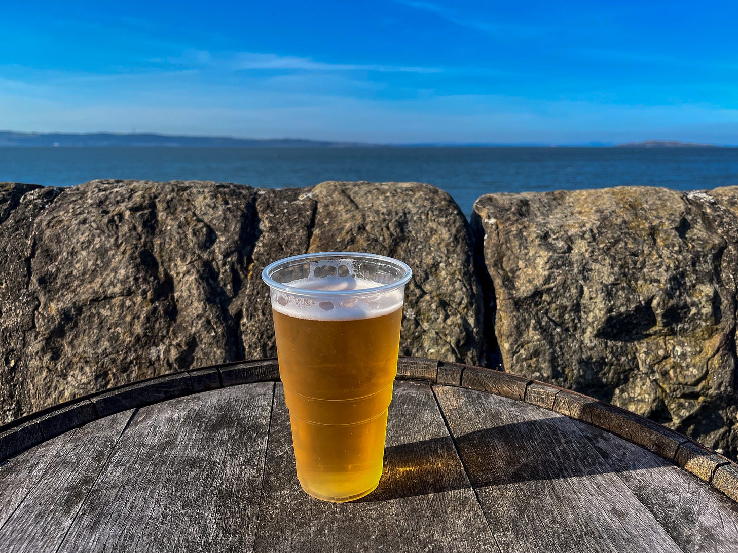 A plastic cup of lager on the top of an old barrel. In the background is the bright blue Firth of Forth.