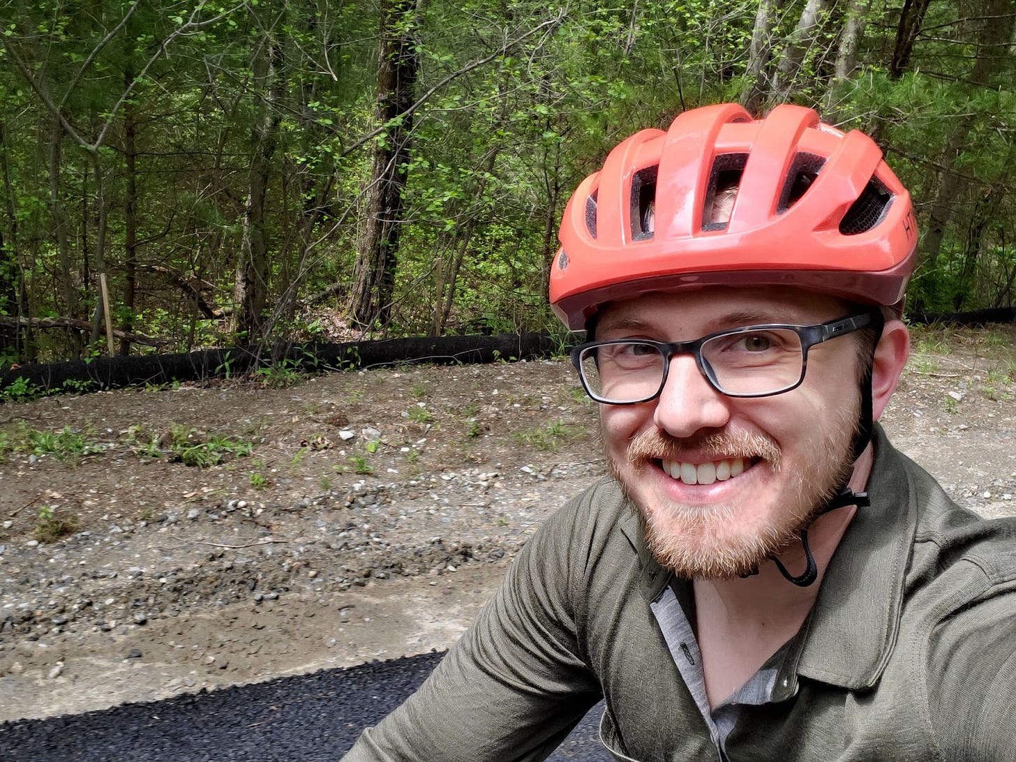 Michael, a white man with a blonde beard, smiles atop a bike. He wears a red helmet and is riding past a forest. Michael, a white man with a blonde beard, smiles atop a bike. He wears a red helmet and is riding past a forest.
