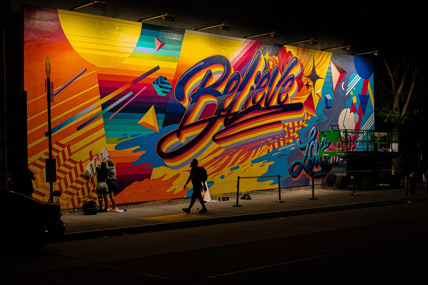 A wall covered in art with Believe in big words. A man walks past while two women work on it further A wall covered in art with Believe in big words. A man walks past while two women work on it further