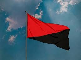 This picture shows a red and black syndicalist flag, a symbol of anarchosocialism, flying from a pole on a partly cloudy day.