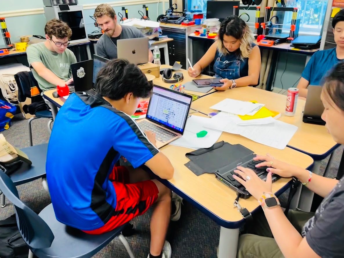 A group of teenagers and an adult sit around a table, working on laptops. A group of teenagers and an adult sit around a table, working on laptops.