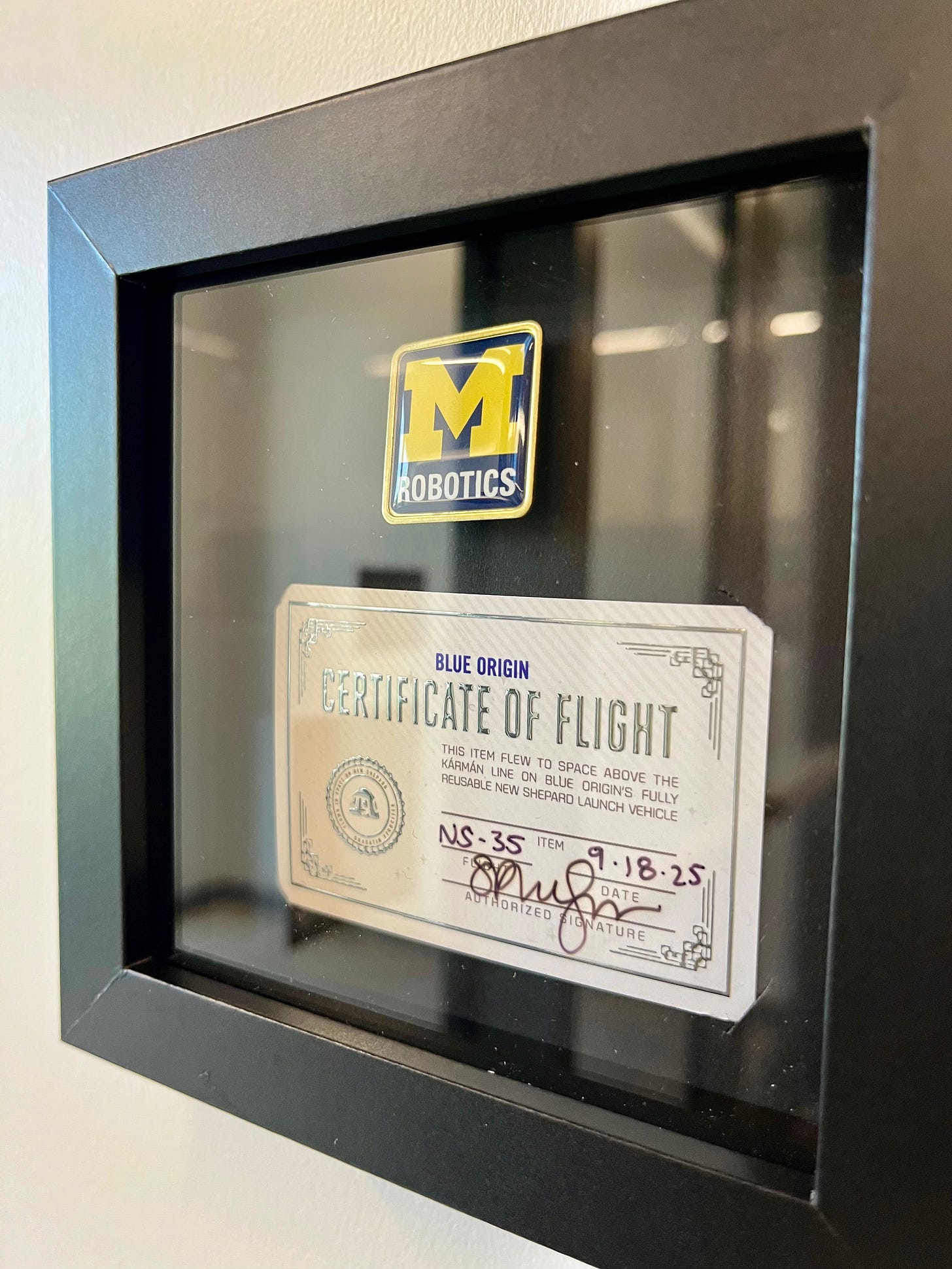 A Michigan Robotics lapel pin sits in a black frame next to a business card-sized certificate of flight from Blue Origin that says this object passed the Karman Line into space on the New Shephard launch vehicle.