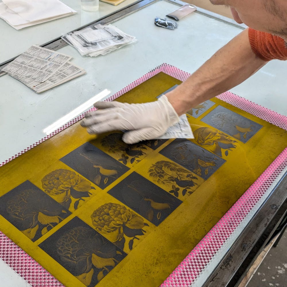 A person wearing a white glove is carefully wiping down a yellow printmaking plate, which features a series of intricate floral and botanical illustrations arranged in a grid. The workspace includes a glass table with tools, newspapers, and a smartphone, suggesting a hands-on printmaking process.