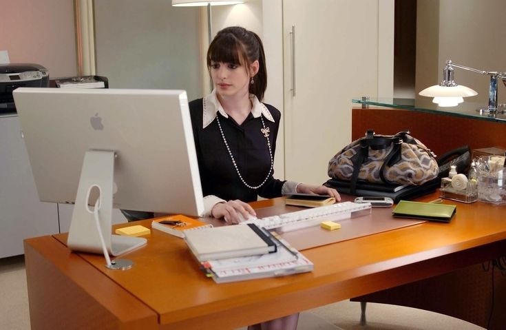 This may contain: a woman sitting in front of a computer on top of a wooden desk next to a purse