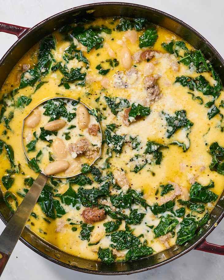 Creamy soup with kale, white beans, and sausage in a red pot, topped with grated cheese and black pepper.