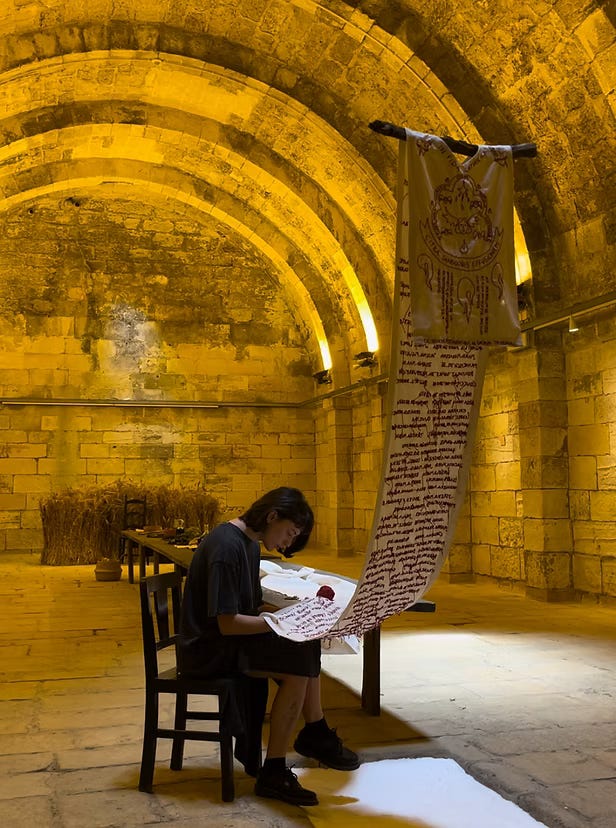 Un'installazione artistica all'interno di una cisterna in pietra, dove una persona seduta ricama con filo rosso su un lungo tessuto bianco, che scende da una struttura in legno. Un'installazione artistica all'interno di una cisterna in pietra, dove una persona seduta ricama con filo rosso su un lungo tessuto bianco, che scende da una struttura in legno.