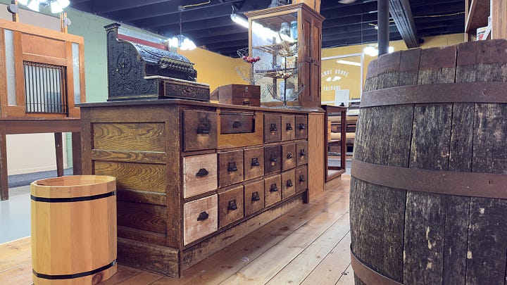 A curly-haired girl picking out cans of vegetables from an interactive mercantile. | Close up of black, lace-up ladies boots. | Low angle from behind the sales counter showing off several cupboards and an old cash register. | Same girl checking mail in the postal cubbies.