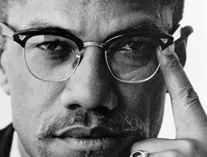 Black and white closeup head shot of Malcolm X looking right at you with a look of extrene gravitas. His left index finger is pointed toward his temple in a "thinking pose".