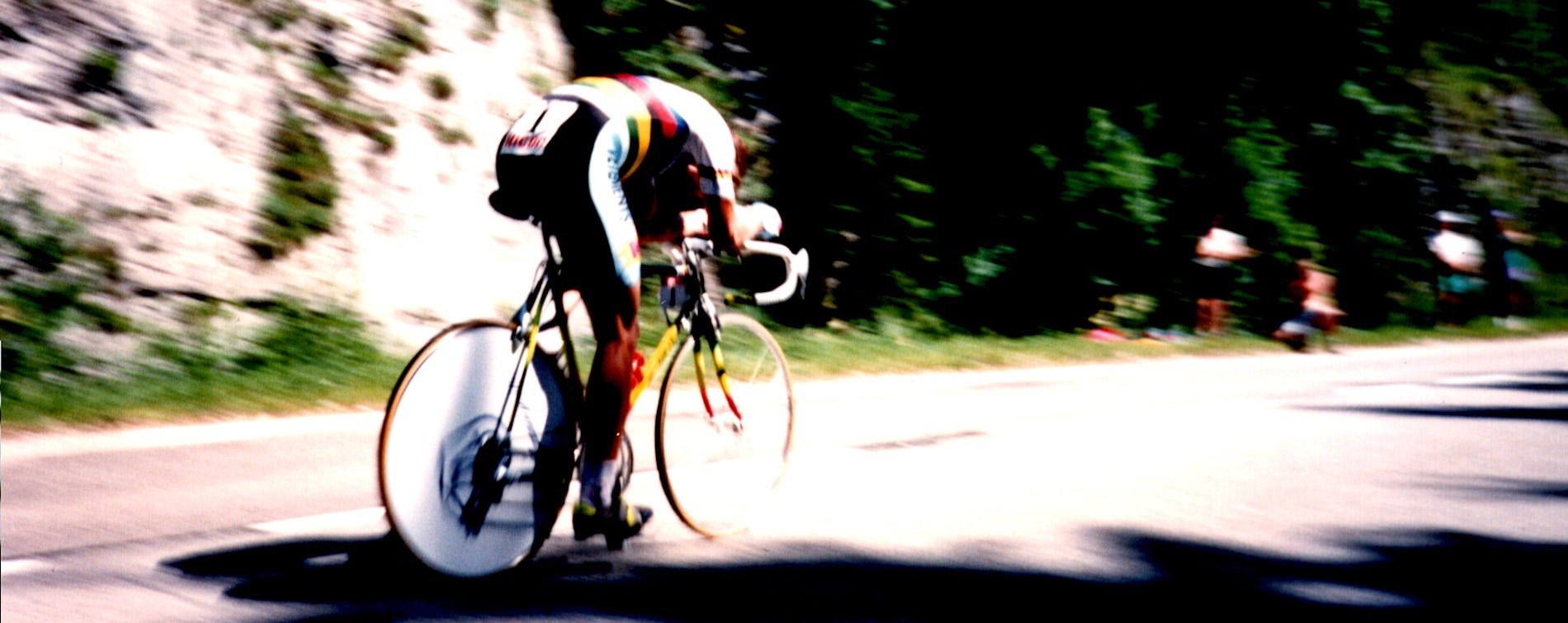 Photo of American bicycle racer Greg Lemond. He is on a racing bike in the mountains. He is pedaling fast - the photo is blurry. His arms are on the aerobars and his head is tucked low for aerodymanics. He is wearing the rainbow jersey of the world champion.