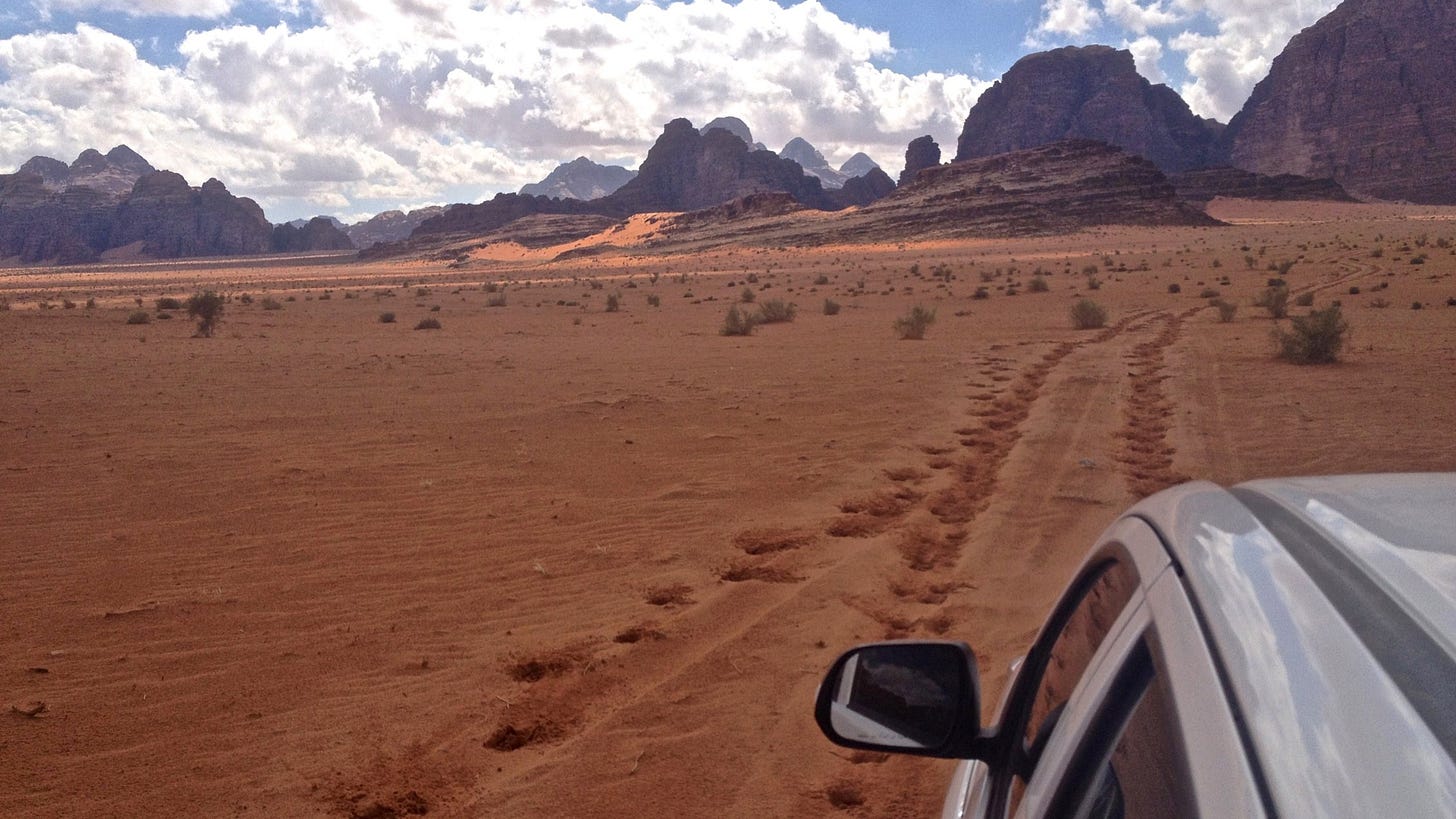 A desert of orange sand, with a trail of camel footprints, leads from the cab of a pick-up truck towards tall, rocky mountains and a blue sky full of bright clouds