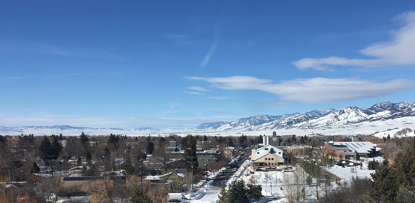 View of Bozeman, Montana from Pete’s Hill in winter, with snow-covered rooftops, distant mountains, and a blue sky overhead.