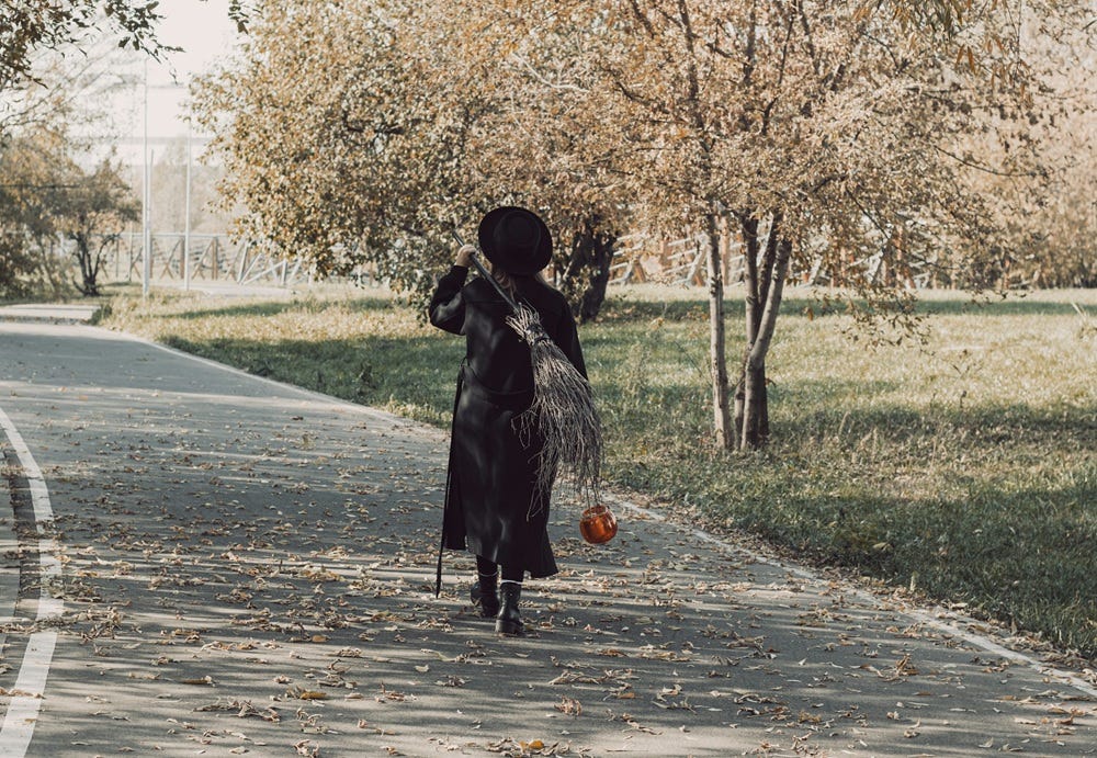 Woman in a witch costume walking through a park holding a pumpkin and with a broom slung over her shoulder.