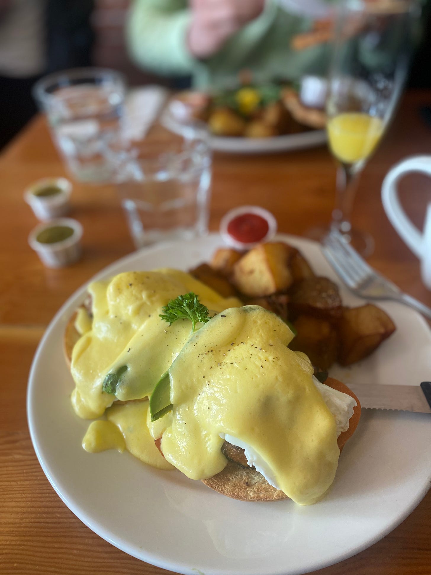 A big plate with eggs, chorizo, tomato, and avocado covered in hollandaise sauce on top of two halves of a bagel. Large hash browns are on the back half of the plate, and a serrated knife is partially wedged beneath one of the bagels. In the background are glasses of water and paper ramekins of ketchup and hot sauce, and a nearly-empty mimosa.
