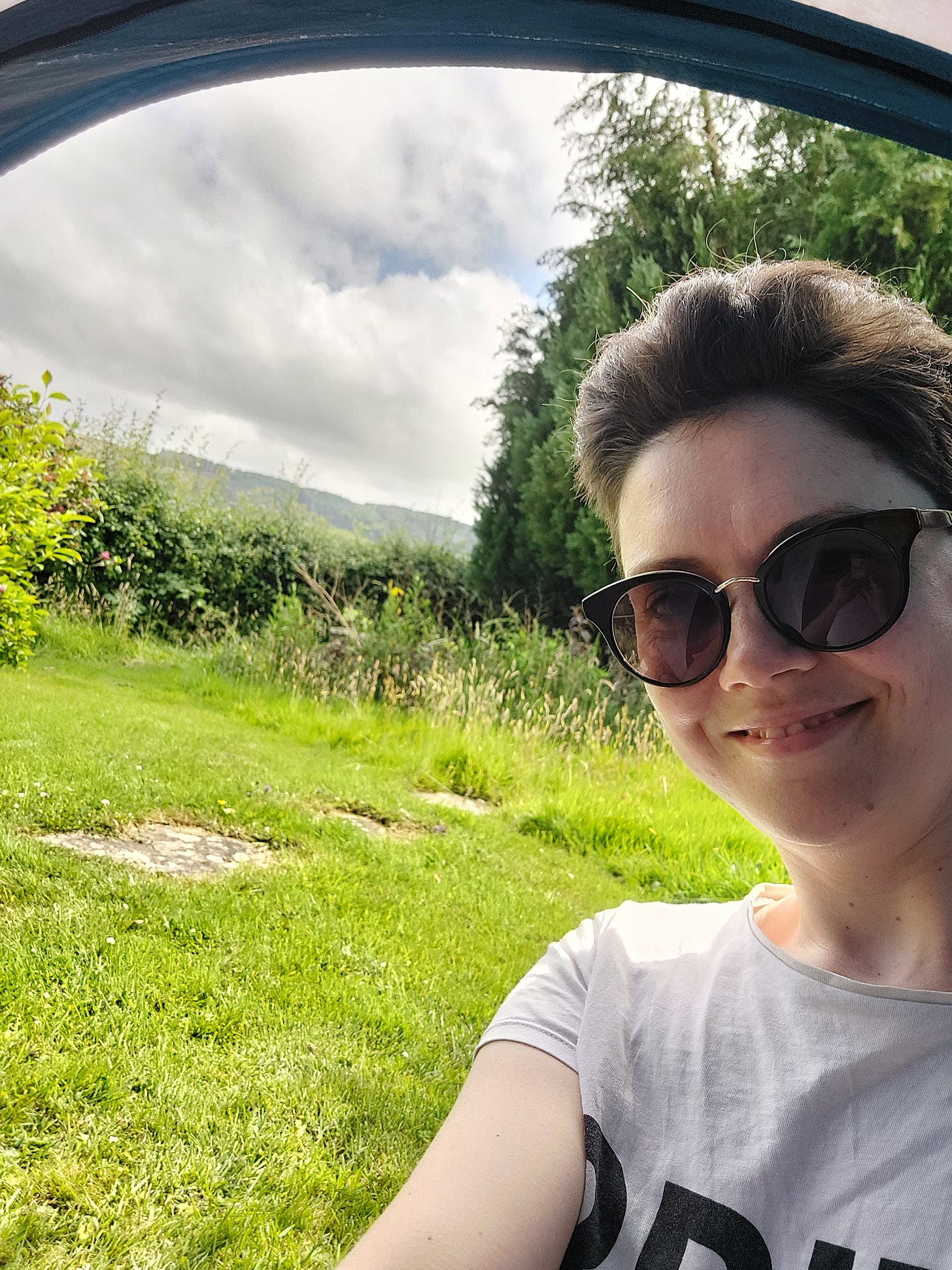 A smiling selfie of Janelle in sunglasses taken from inside a tent. Outside is green grass, foliage and a grey cloudy sky