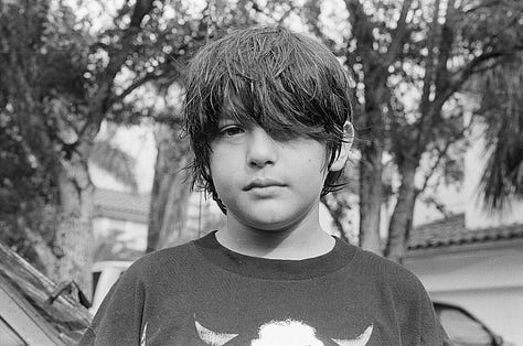 Counter-clockwise from top left: view from photographer's eye-level perspective of her feet in converse sneakers; three customers framed by two cars in adjacent parking lot at a ventanita, or coffee window, of a Cuban restaurant; portrait of young boy looking straight to camera with trees in background; image of photography lab; skyward view of sun poking through oak trees with and American flag in opposite corner; landscape view of a strip mall in suburban Miami.