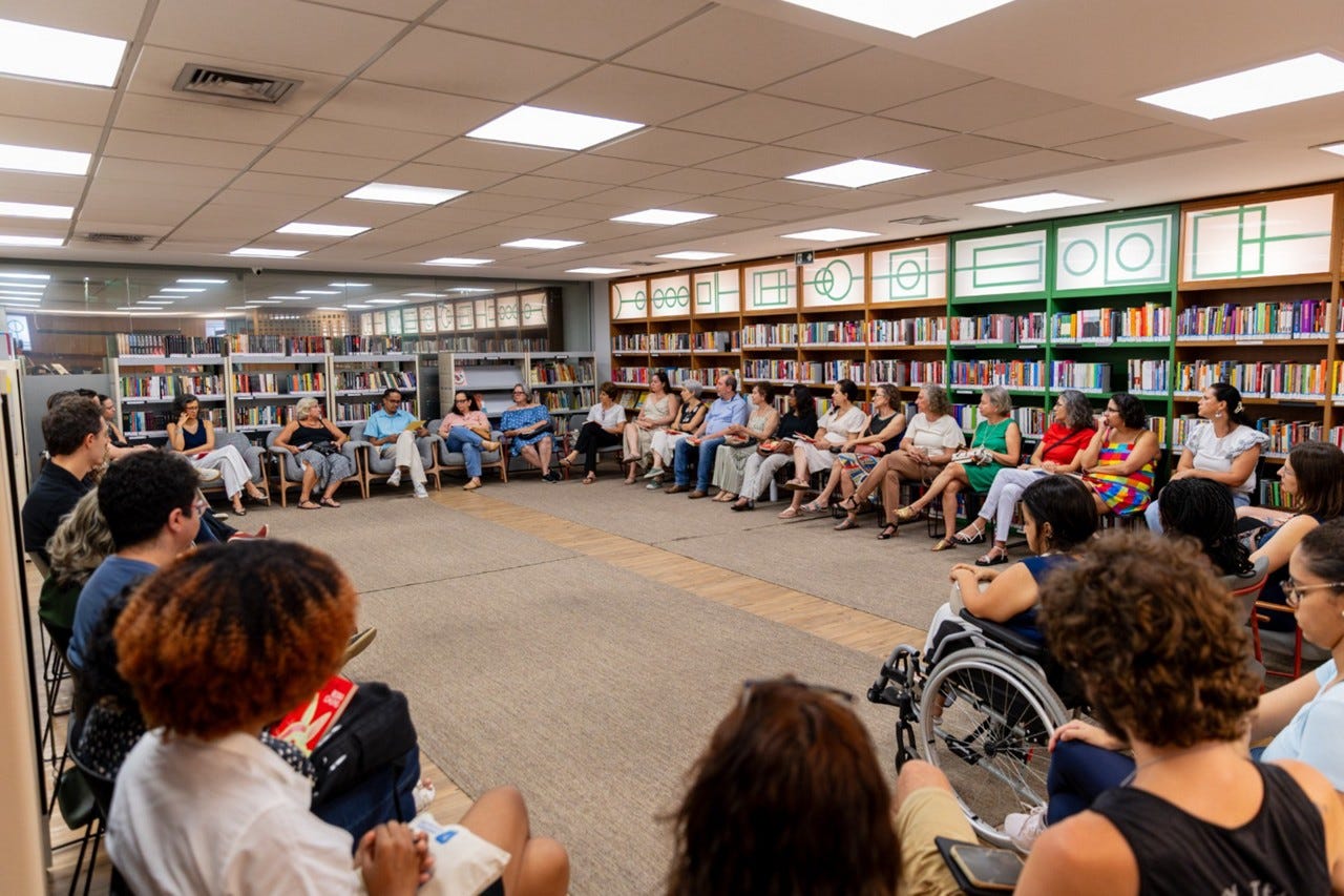 #descrevemospravocê  Fotografia de um encontro realizado em uma biblioteca. Cerca de trinta pessoas estão sentadas em círculo, ocupando todo o espaço da imagem. As cadeiras estão dispostas ao redor de um grande tapete bege, que cobre o centro do salão. Ao fundo, prateleiras repletas de livros ocupam toda a parede, e acima delas há painéis com molduras verdes e formas geométricas em branco. O teto é de placas retangulares com luminárias embutidas que iluminam bem o ambiente.  O grupo é diverso, formado por homens e mulheres de diferentes idades e estilos, com variações de tons de pele, roupas e posturas. Entre os participantes, há uma mulher usando cadeira de rodas e pessoas segurando livros ou cadernos. Algumas pessoas estão de pernas cruzadas, outras com as mãos no colo ou gesticulando, em um clima de escuta e troca. O ambiente é acolhedor e organizado, transmitindo a ideia de uma roda de conversa, grupo de leitura ou encontro comunitário.
