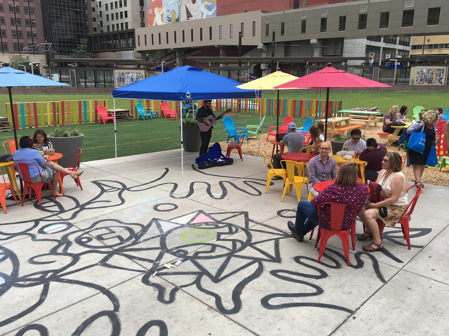 People sit under colorful umbrellas at tables at a downtown transit stop.