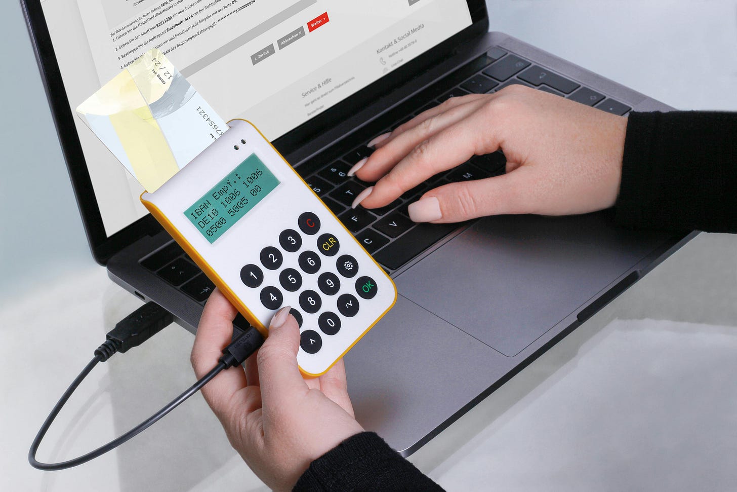 A close-up, high-angle shot of a person’s hands using a laptop and a handheld chip-and-pin card reader. A bank card is inserted into the white and yellow device, which displays an IBAN number on a green digital screen. The person's other hand is poised over the laptop keyboard, suggesting an active online banking transaction. A close-up, high-angle shot of a person’s hands using a laptop and a handheld chip-and-pin card reader. A bank card is inserted into the white and yellow device, which displays an IBAN number on a green digital screen. The person's other hand is poised over the laptop keyboard, suggesting an active online banking transaction.