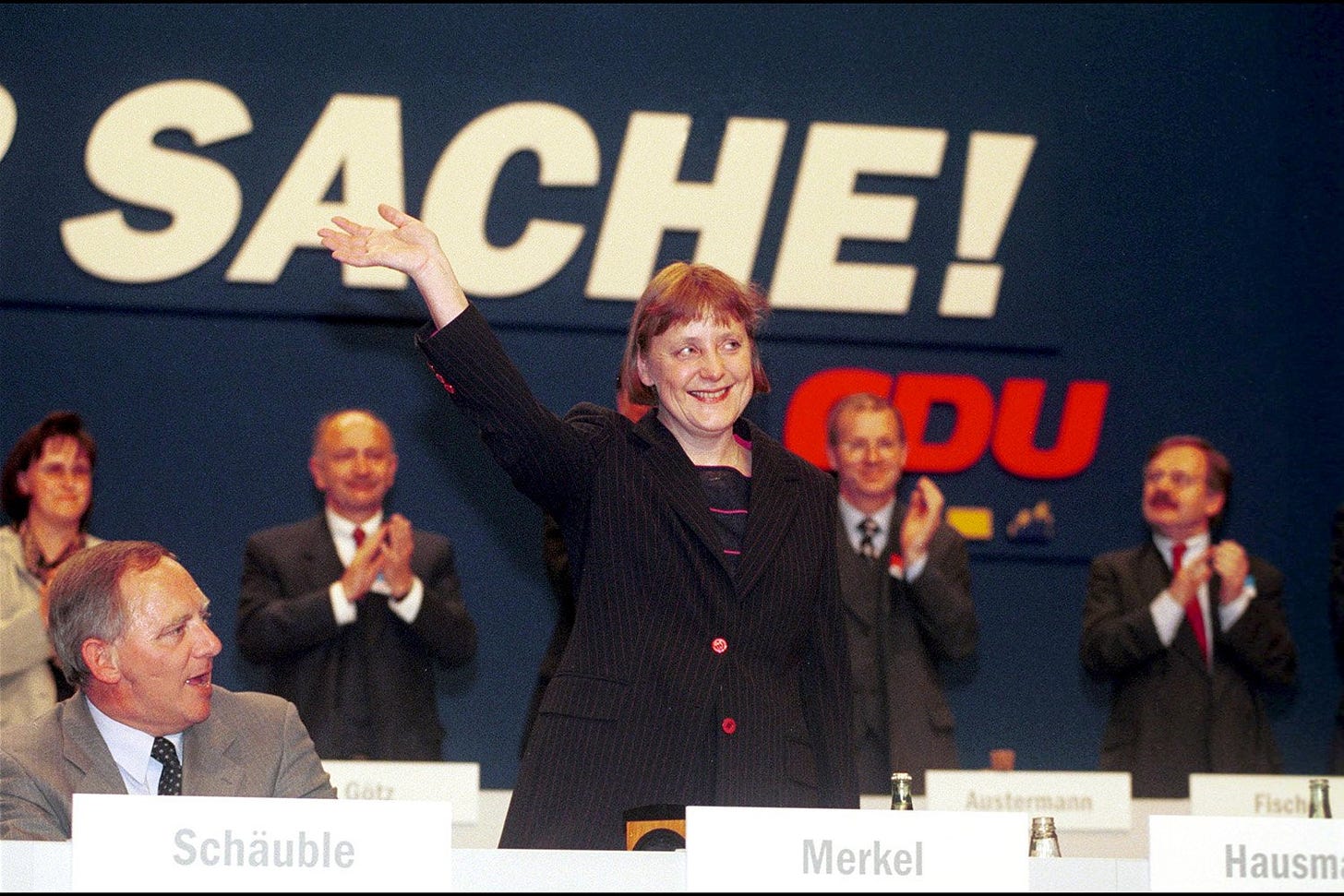 A vintage photograph of Angela Merkel early in her career, standing to wave and smile at an audience after an election. Her hair is slightly longer than in the other photos, and she wears a black pinstripe blazer with pink buttons.
