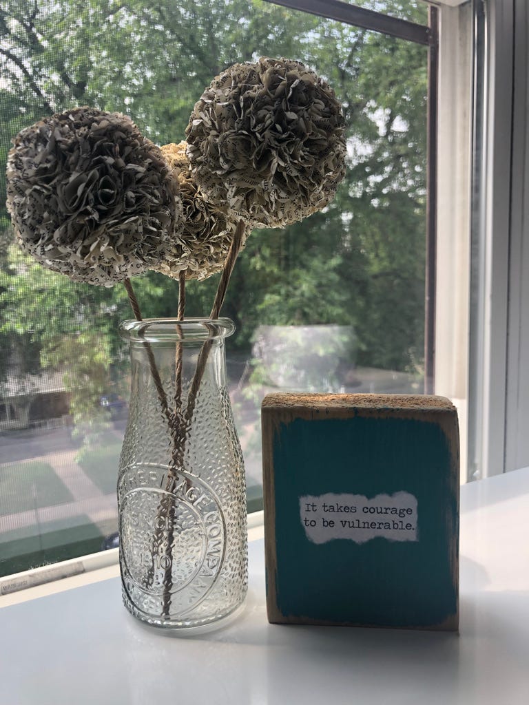 Glass vase with 3 hydrangea-like blossoms made of paper, and a small teal colored sign that reads "It takes courage to be vulnerable." Glass vase with 3 hydrangea-like blossoms made of paper, and a small teal colored sign that reads "It takes courage to be vulnerable."