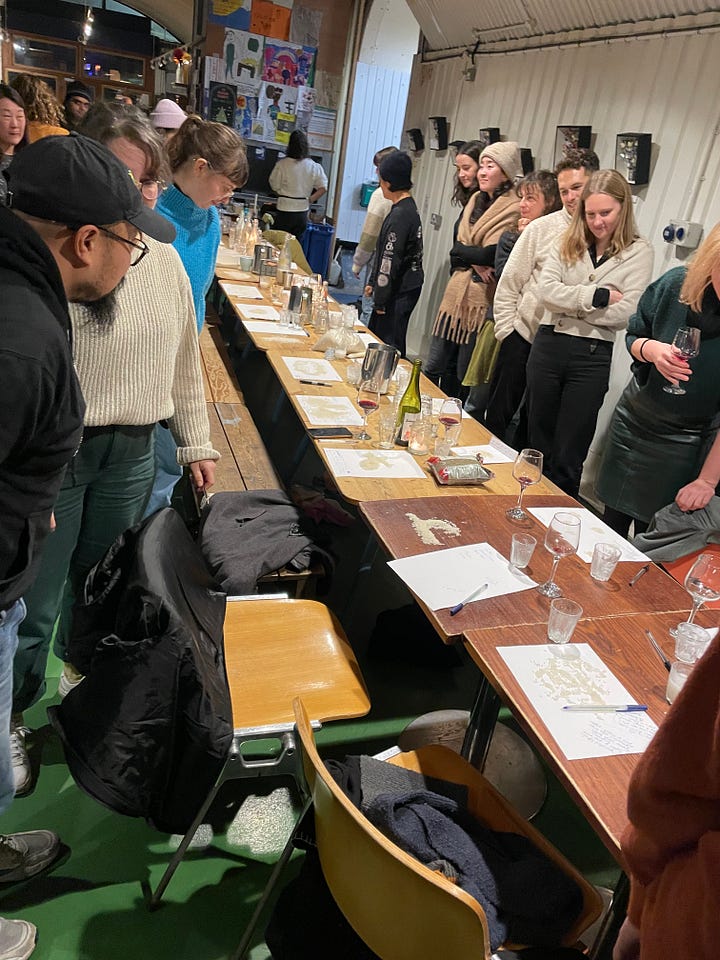 The first image is of a long wooden table with different bags of rice in an otherwise empty railroad arch. The second image is the same table with people walking around and examining eachothers rice artwork