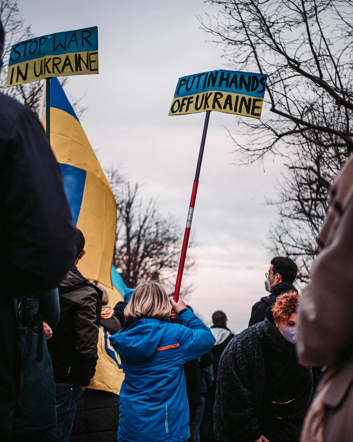 A young girl at a demonstration holds a sign that says "Putin Hands Off Ukraine." A nearby sign says "Stop War In Ukraine."