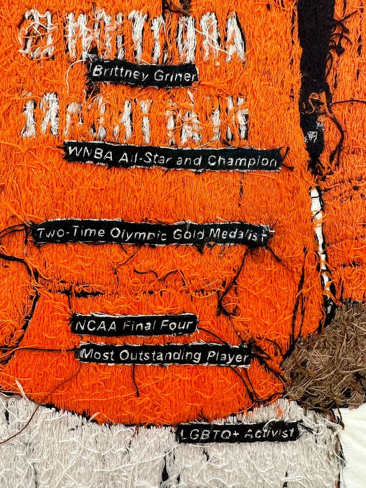 1 - 2) Brittney Griner's portrait is seen fron the front view. In it, she is wearing an orange hoodie that says 'Abortion is healthcare'. She has dreads, some of which are tucked into her hoddie, and she is wearing white shorts, and white leggings underneath. She has on black sneakers, and black backpack straps on her shoulders. She also has thin-framed black square glasses on. 3) The verso side of the embroidery is seen with different messages attached, and messy stringwork. 4) A close-up of the verso side shows some of the texts strings on black fabric like 'Brittney Griner', 'Two-time olympic gold medalist', and 'NCAA Final Four'. 5) The front of the portrait is seen at a gallery, surrounded by two other works on eithe side. It's sitting on a tall white pedestal. The gallery's walls are white, with black stone flooring.