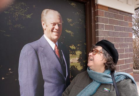 Annette with a lifesize mural of President Gerald Ford; Pulling a small covered cart on two wheels; with two kids in a museum atrium; With staues of Franklin and Eleanor Roosevelt sitting at tables; taking picture of photo exhibit; Outdoors with monuments and road in background.