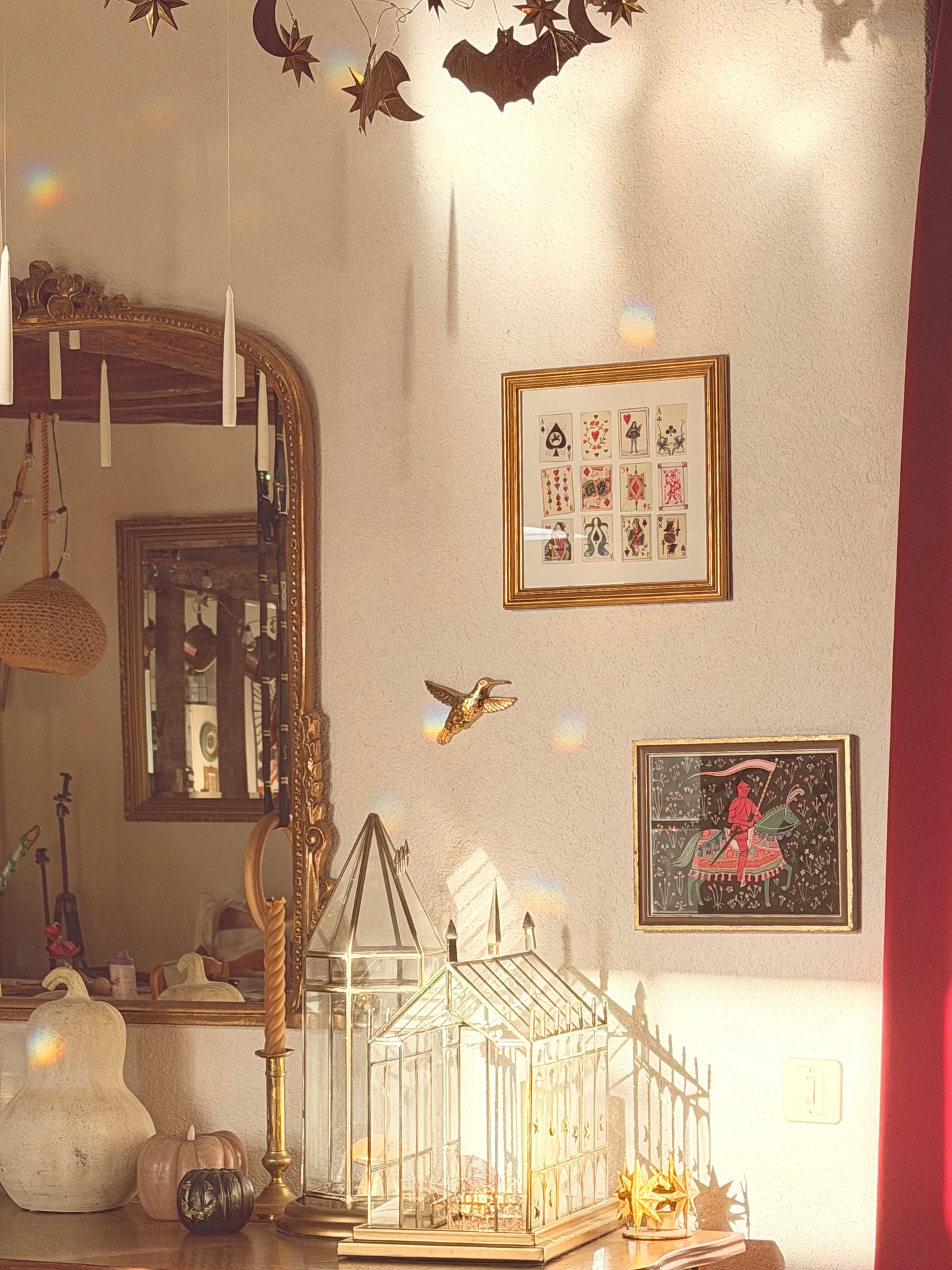 A sunlit corner of a château room decorated for Halloween. Paper bats, moons and stars hang from the ceiling above a row of floating white taper candles. Below, there’s a gilt arched mirror, a brass hummingbird on the wall, framed antique playing cards and a painting of a red knight on horseback. On the sideboard sit two glass and brass terrarium houses lit with fairy lights, a brass candlestick, a tall pale gourd and small pink and black pumpkins, with little rainbow reflections scattered across the wall. A sunlit corner of a château room decorated for Halloween. Paper bats, moons and stars hang from the ceiling above a row of floating white taper candles. Below, there’s a gilt arched mirror, a brass hummingbird on the wall, framed antique playing cards and a painting of a red knight on horseback. On the sideboard sit two glass and brass terrarium houses lit with fairy lights, a brass candlestick, a tall pale gourd and small pink and black pumpkins, with little rainbow reflections scattered across the wall.