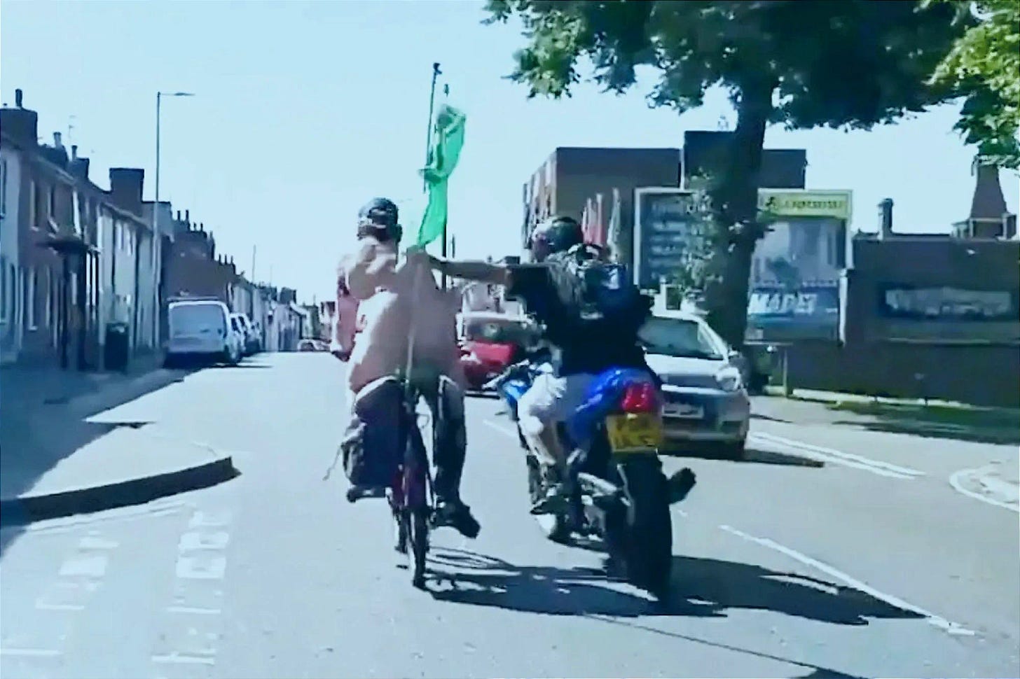 A still image from street video shows a nude cyclist riding on a sunlit residential road as a motorcyclist pulls alongside and reaches across with an outstretched arm. Parked cars line the street and a green flag is mounted behind the cyclist.