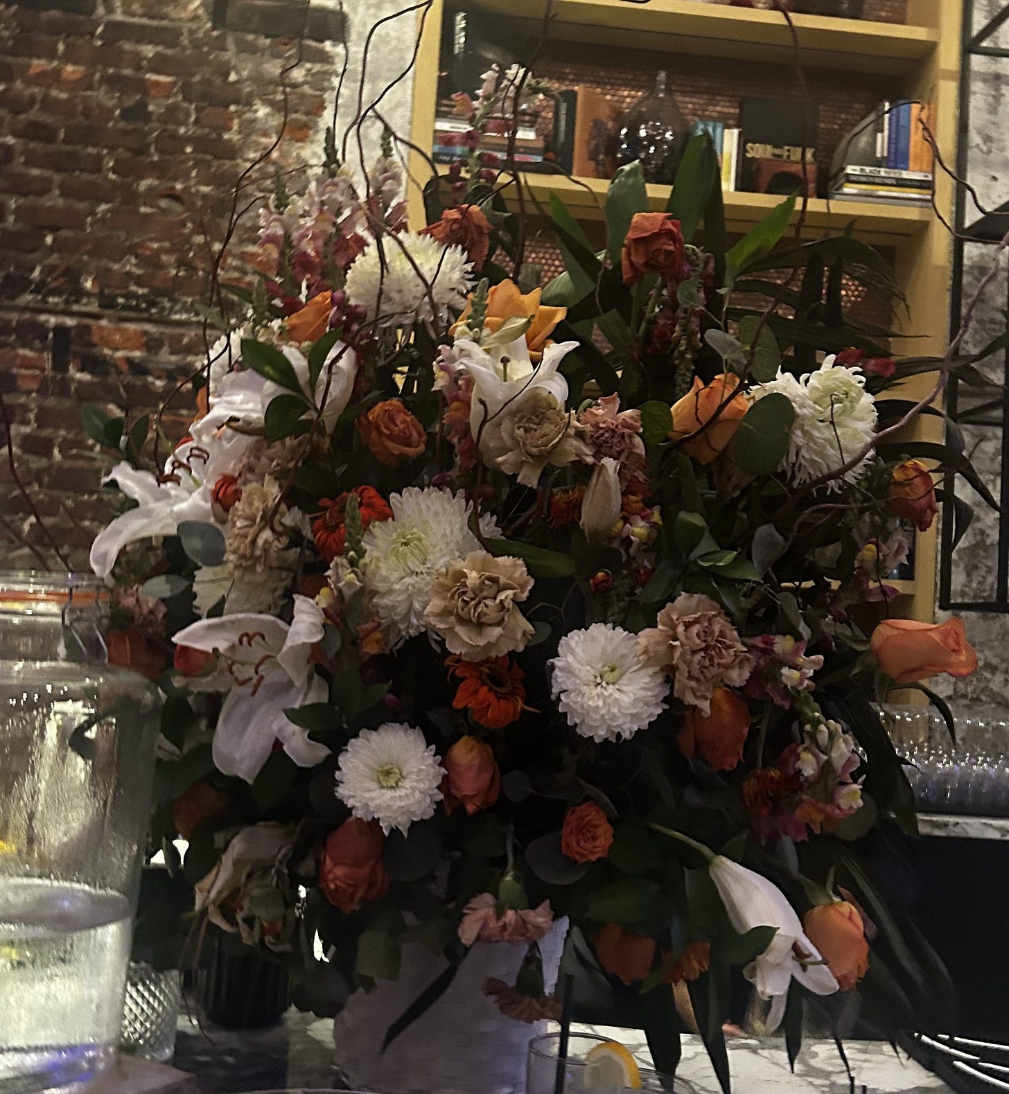 Lush floral arrangement with roses, lilies, and mixed blooms in warm autumnal tones displayed on a bar counter. Soft ambient lighting highlights the layered textures of petals and greenery, creating a romantic still-life scene against a brick wall and bookshelf backdrop. Lush floral arrangement with roses, lilies, and mixed blooms in warm autumnal tones displayed on a bar counter. Soft ambient lighting highlights the layered textures of petals and greenery, creating a romantic still-life scene against a brick wall and bookshelf backdrop.